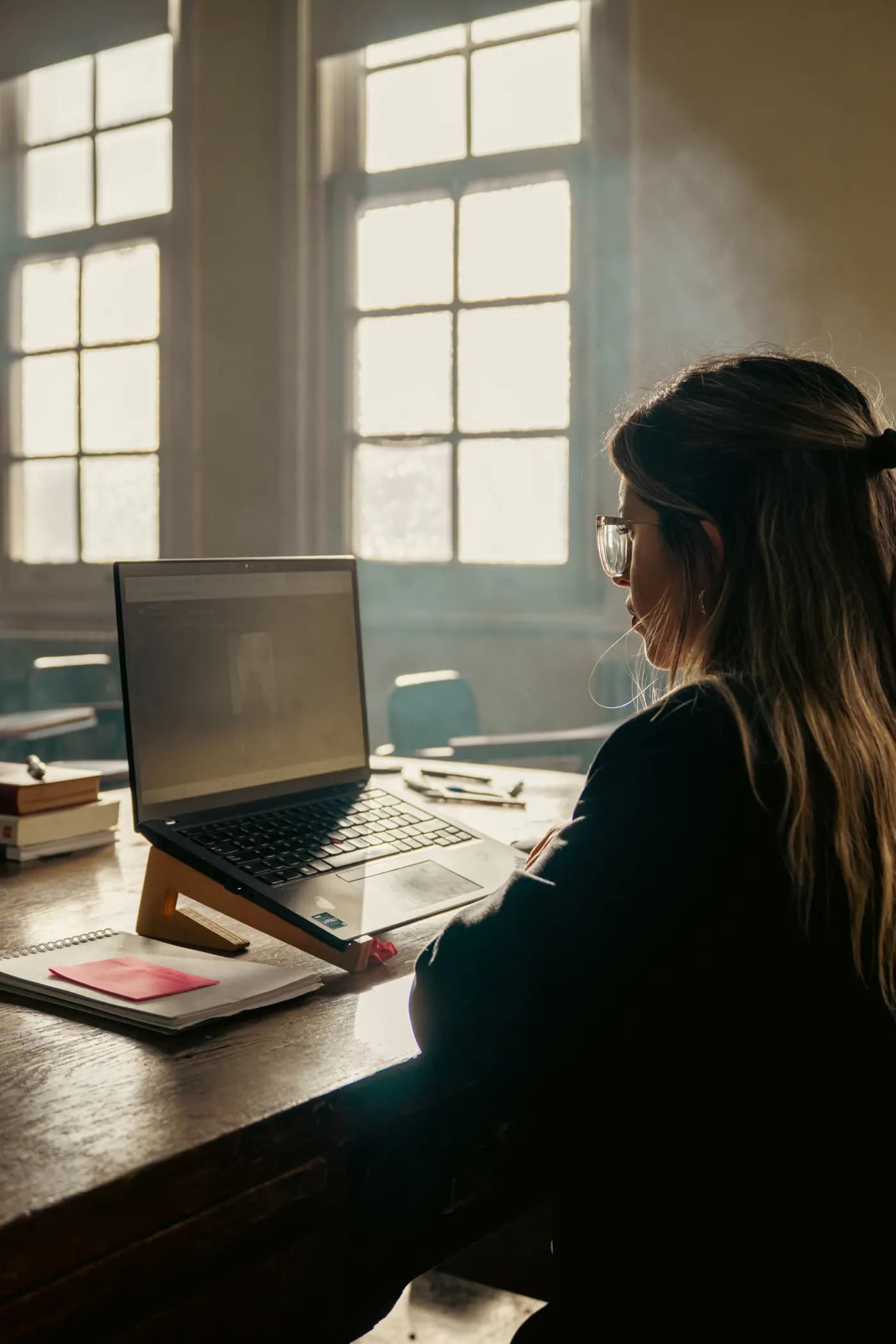 A student seen from behind working at their laptop in a classroom, daylight through tall windows, a notebook and books on the desk