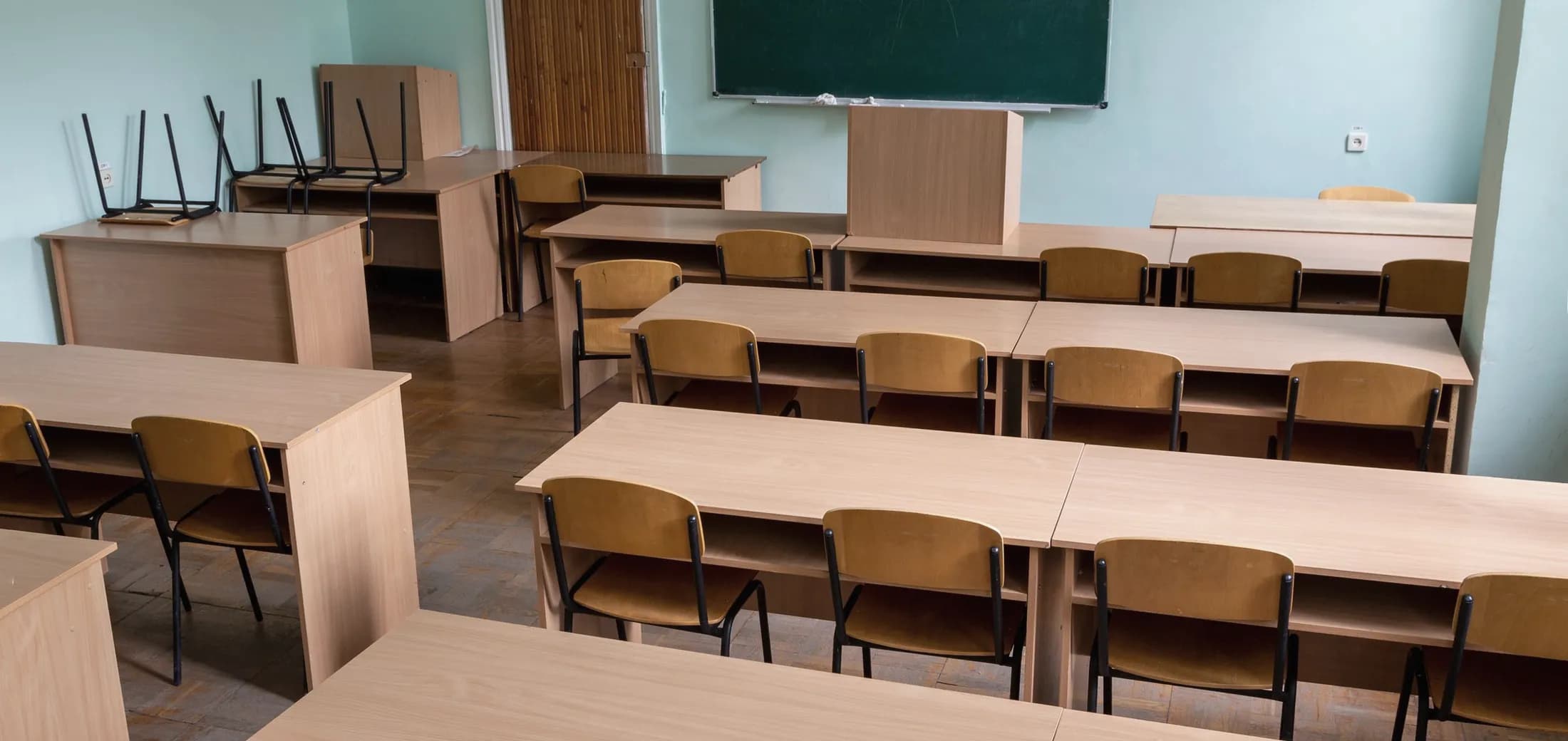An empty classroom, rows of light wooden desks, a green chalkboard and chairs stacked on the tables at the back