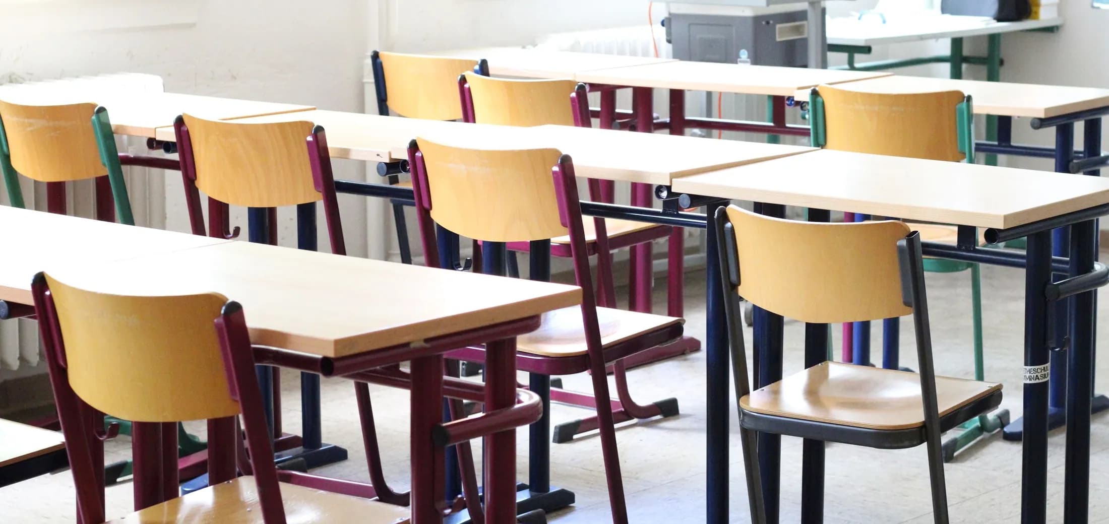 A bright, empty teaching room, rows of pale wooden desks and chairs, nobody present